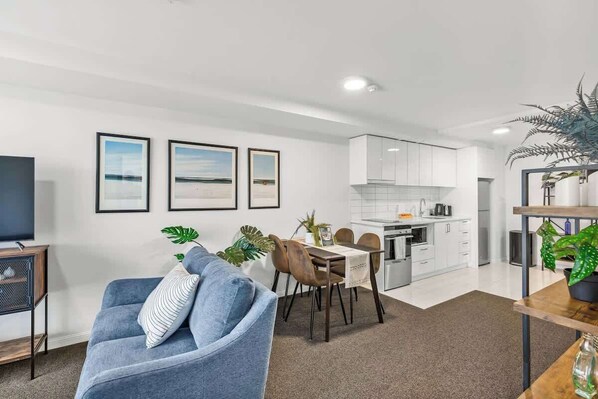 A modern living room with a blue sofa, a dining area with a table and chairs, a kitchen with white cabinets, and framed pictures on the wall.