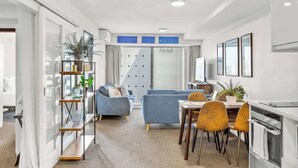 Una sala moderna con sofá, mesa de comedor y sillas, una zona de cocina con horno, y un ventanal con cortinas.