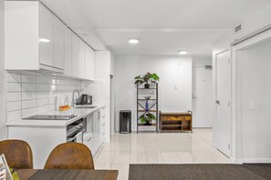 A modern kitchen with white cabinets, a built-in oven, and a coffee machine. There is a dining area with a table and chairs, and a small plant on a shelf. - 2BR Apt w/ Parking in Prime Spot by Zodiak Stays (Auckland)