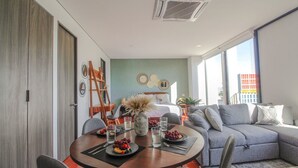 A modern living room with a dining table set for two, a grey sofa, and a large window providing a view of the cityscape. - Departamento exclusivo en Chapultepec 67 (Guadalajara)