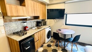 Ett kompakt kök med gasspis, mikrovågsugn och tvättmaskin, ett litet matbord med stolar och en tv som är monterad på väggen.
