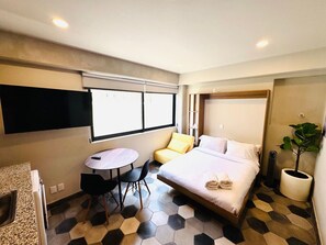 Een hotelkamer met een bed, een kleine tafel met stoelen, een aan de muur bevestigde televisie en een potplant.