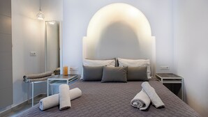 A modern hotel room with a large bed, a bedside table, a nightstand, a mirror, and a unique arched ceiling light fixture. - Santorini Garden Homes (Santorini)