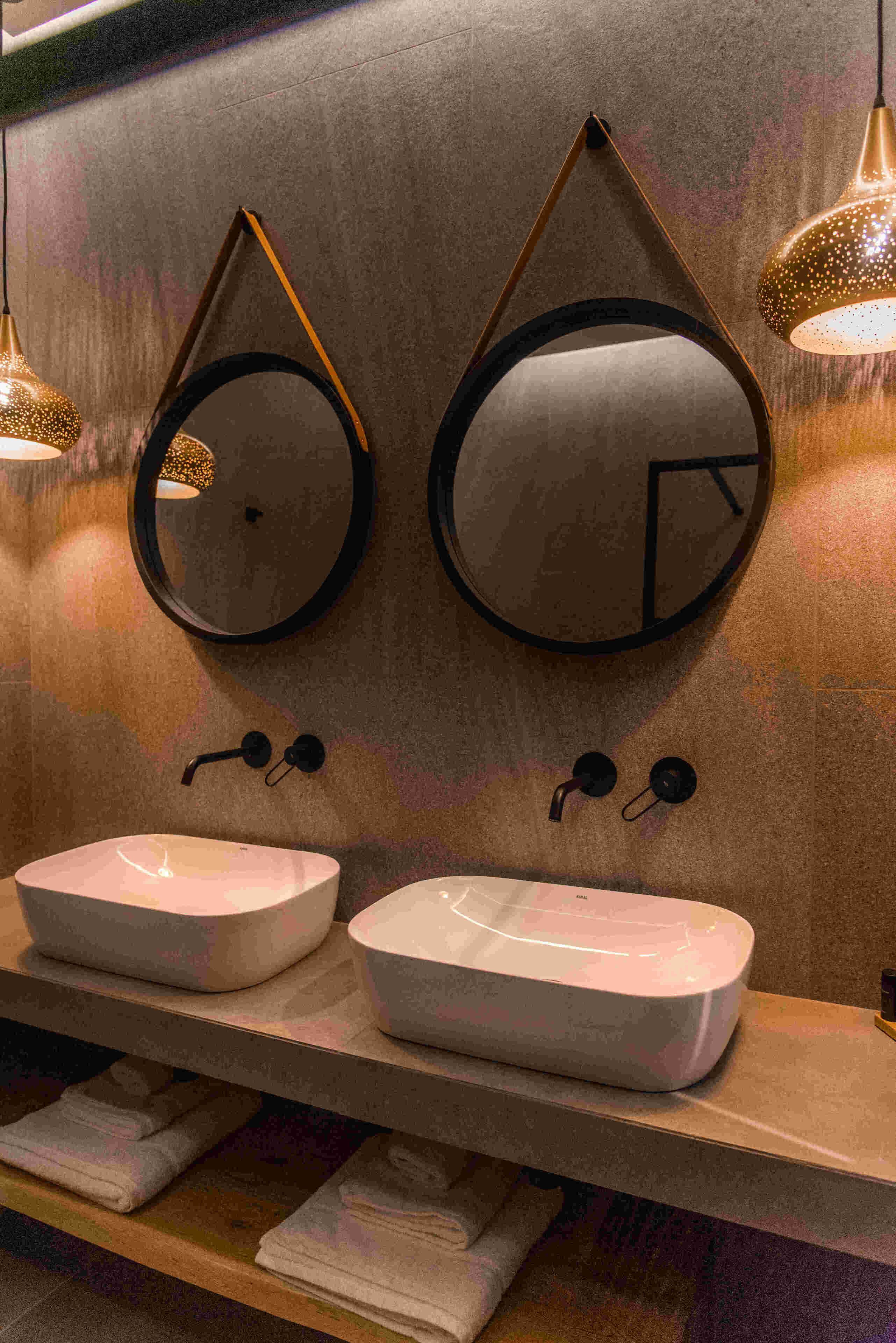 A bathroom with two round mirrors, a double sink with a wooden countertop, and a shelf with folded towels.