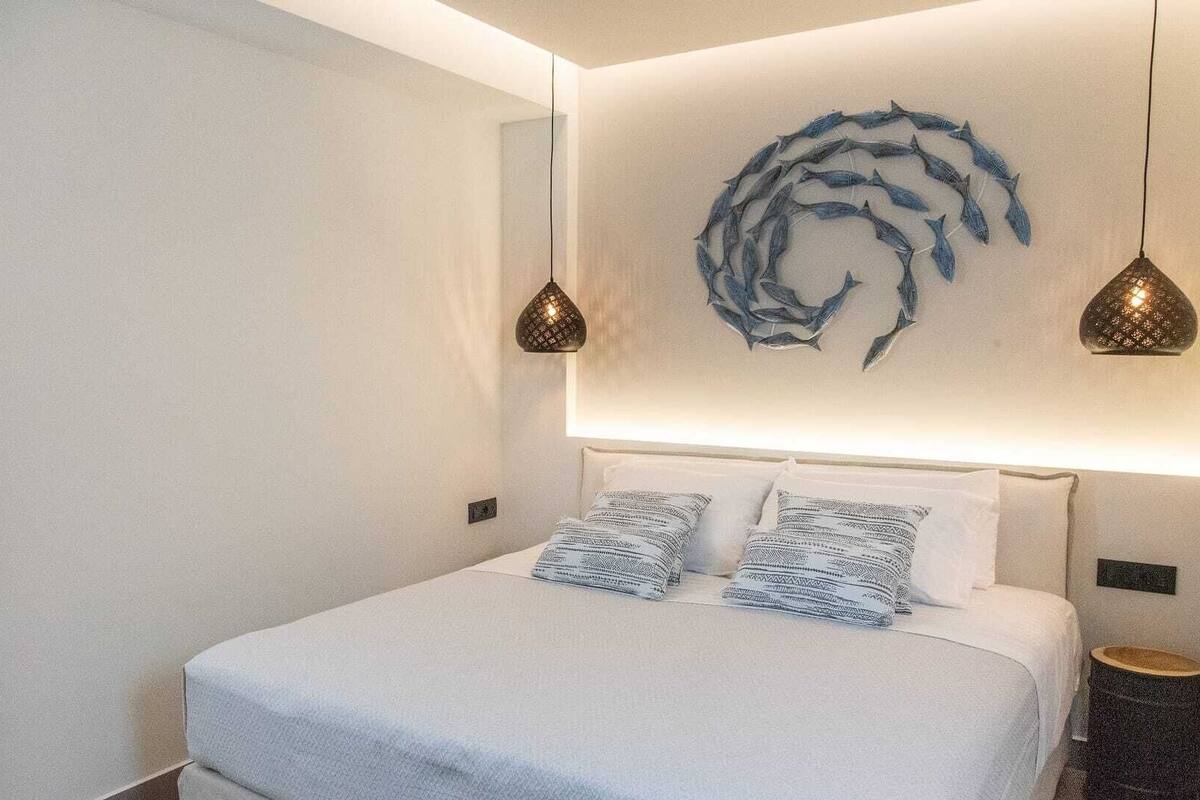A bedroom with a bed, two patterned pillows, a wall art piece, and two pendant lights.