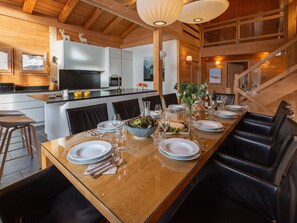 Une cuisine moderne avec des poutres en bois, un coin repas avec une longue table dressée pour six personnes, et un escalier.