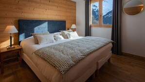 A bedroom with a wooden headboard, a large bed with patterned bedding, two bedside tables with lamps, a window with a view of mountains, and a round mirror on the wall. - 4★ Chalet, 12 pers, spa, panoramic view, near shuttle (La Clusaz)