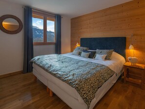 A bedroom with a wooden headboard, a large bed with patterned bedding, a round mirror, and a window with curtains.