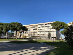 Un edificio residencial de varios pisos con balcones, rodeado de árboles y una zona pavimentada.