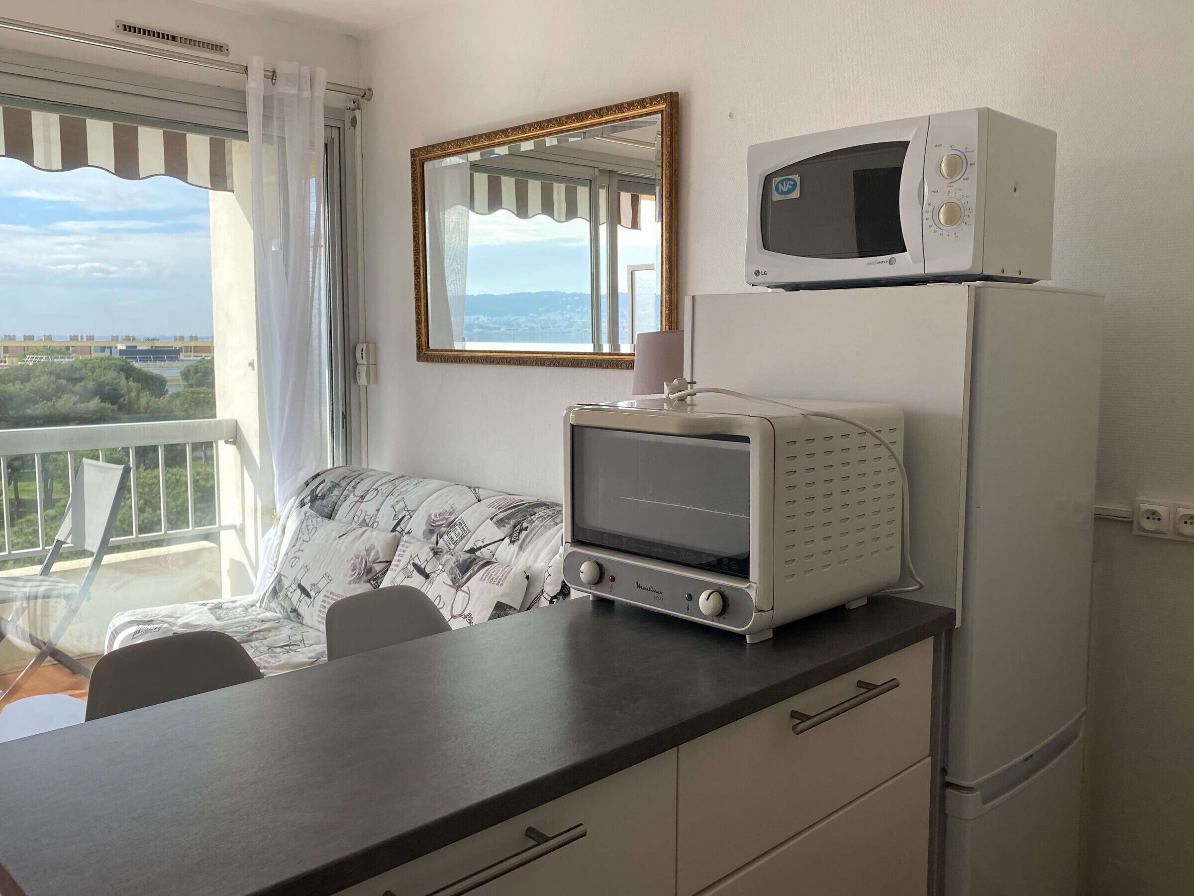 Une pièce comprenant un coin cuisine, un espace repas avec une table et des chaises à motifs, une grande fenêtre offrant une vue et un miroir sur le mur.