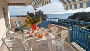 A balcony with a table set for four, surrounded by white plastic chairs, a potted plant, and a glass jar with fruit.