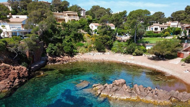 A coastal area with clear waters, rocky shore, and buildings in the background.