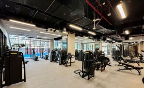Un gimnasio con diversas máquinas de ejercicio, amplias ventanas y un techo alto con tuberías a la vista.