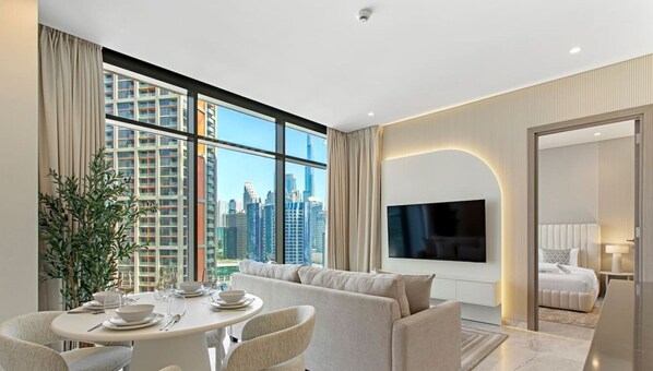 A modern hotel room with a large window offering a city view, a flat-screen TV, a sofa, and a dining area with a table set for two.