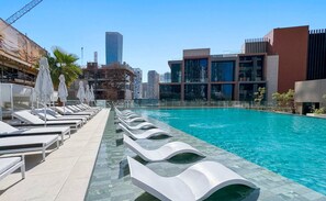 Una piscina con sillas de descanso y sombrillas, rodeada de edificios modernos y un cielo despejado.