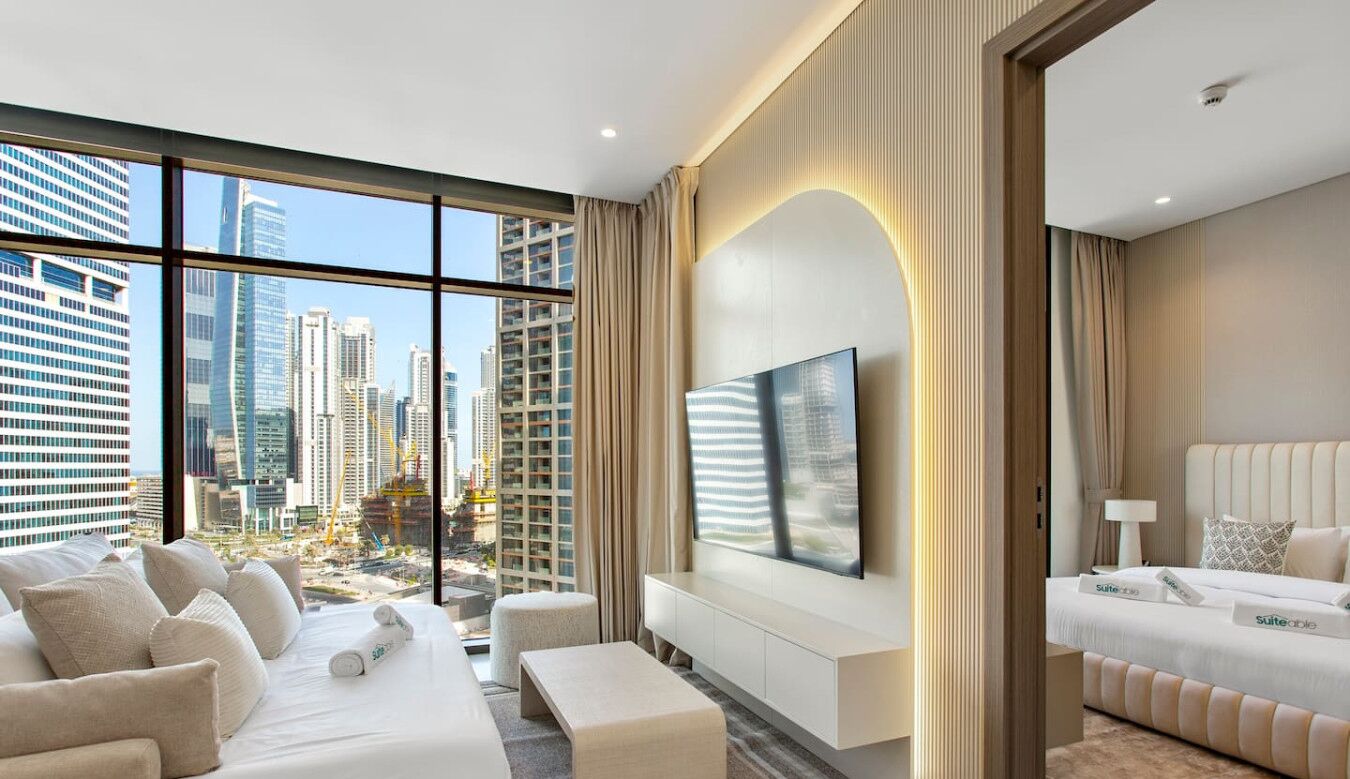 A modern hotel room with a large window, a flat-screen TV, a sofa, a bed, and a view of the cityscape.