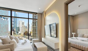 A modern hotel room with a large window, a flat-screen TV, a sofa, a bed, and a view of the cityscape.