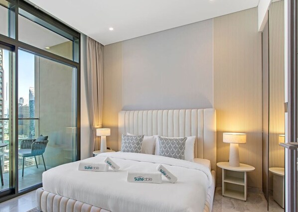 Habitación de hotel con una cama grande, dos mesitas de noche con lámparas, vistas de la ciudad a través de ventanas de gran altura y un balcón con una silla.