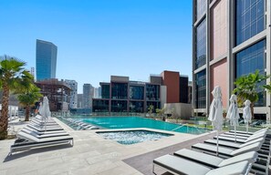 A rooftop pool area with lounge chairs and umbrellas, surrounded by modern buildings and palm trees.