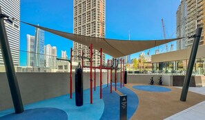 Un gimnasio al aire libre con equipamiento de ejercicio, una zona con sombra y vistas a la ciudad de fondo.