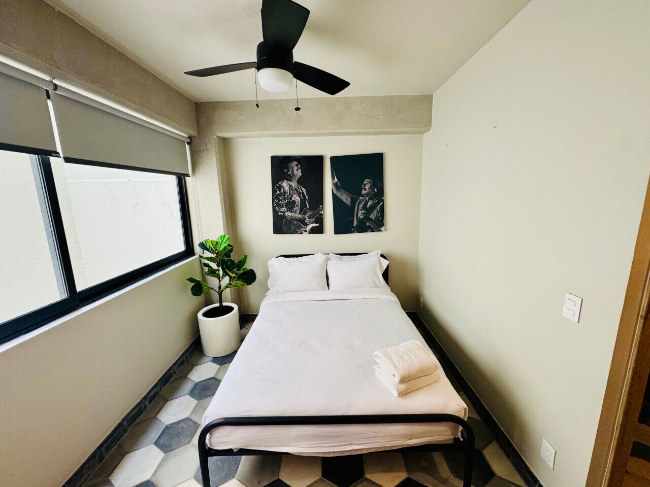 A bedroom with a bed, a ceiling fan, a window with blinds, a potted plant, and two framed pictures on the wall.