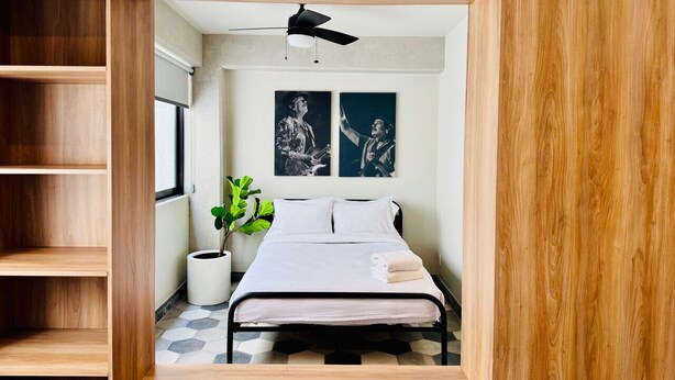Un dormitorio con una cama, un ventilador de techo, dos obras de arte colgadas en la pared, una planta en maceta y una ventana.