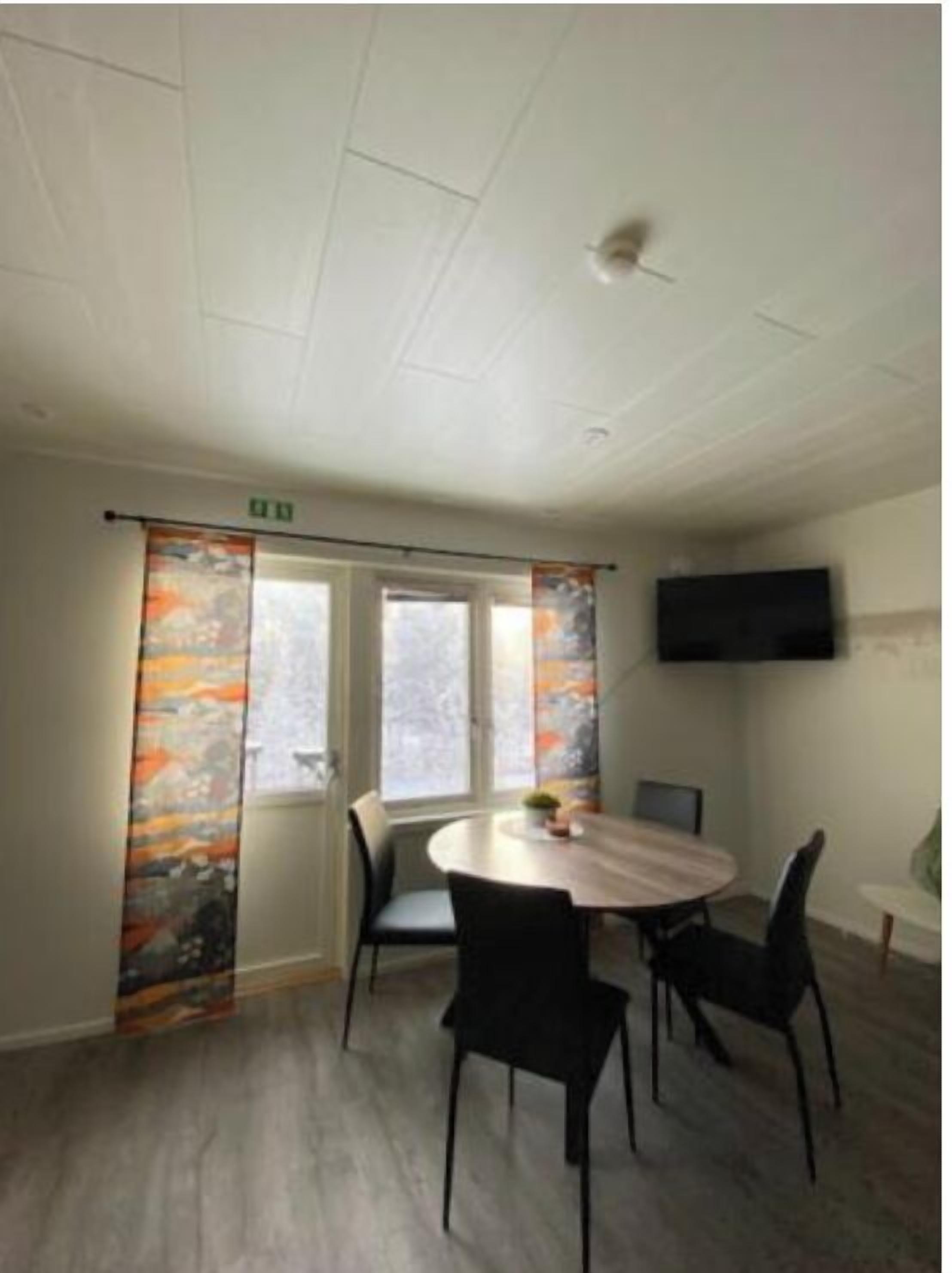 A conference room with a round table, four chairs, a flat-screen TV on the wall, and two windows with curtains.