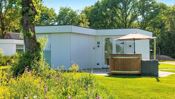 Un bâtiment moderne doté d’une grande porte vitrée, entouré d’un jardin bien entretenu où poussent différentes espèces de fleurs et où se trouve un banc en bois.