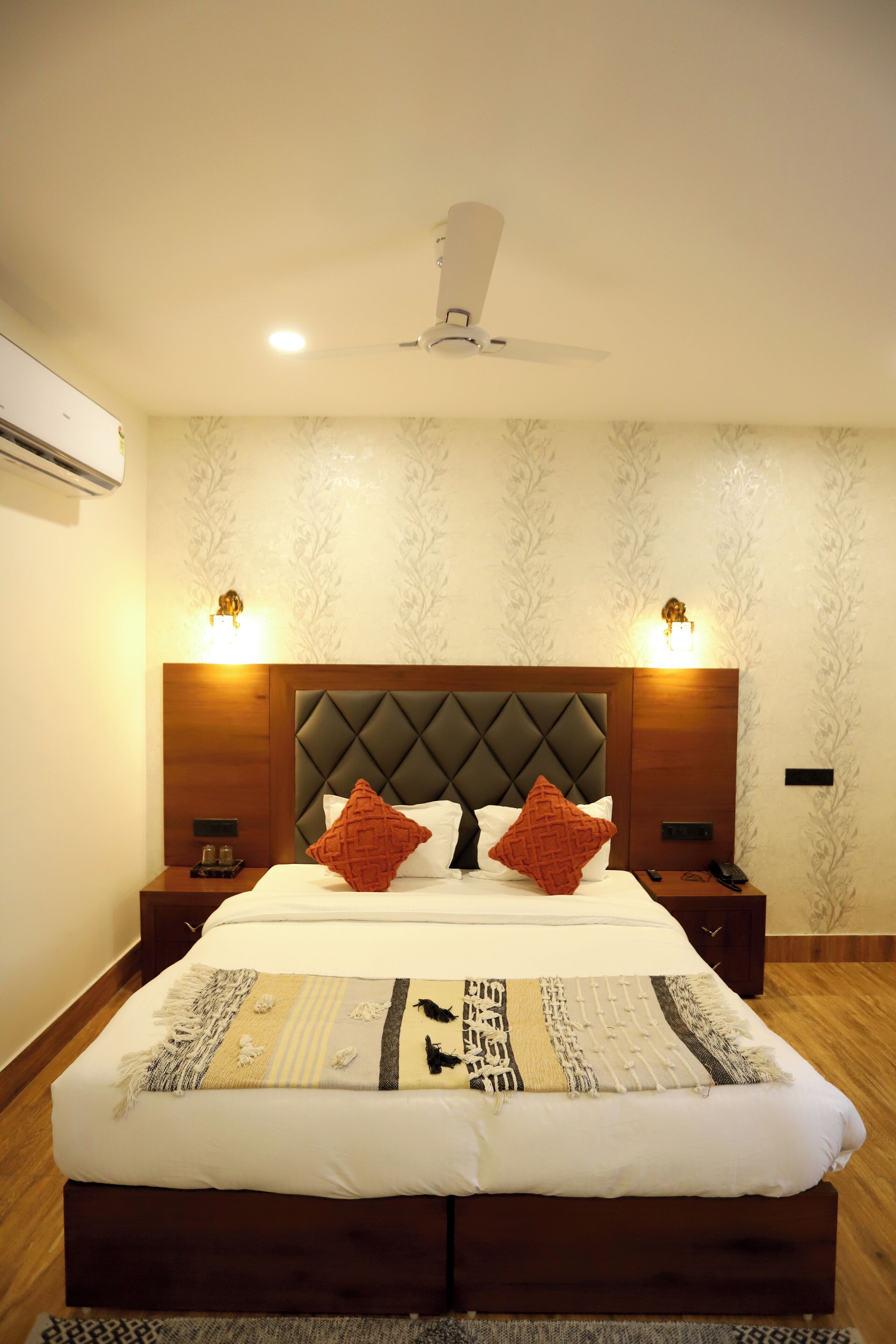 A neatly made bed with a quilt, two bedside tables with lamps, a ceiling fan, and patterned wallpaper.
