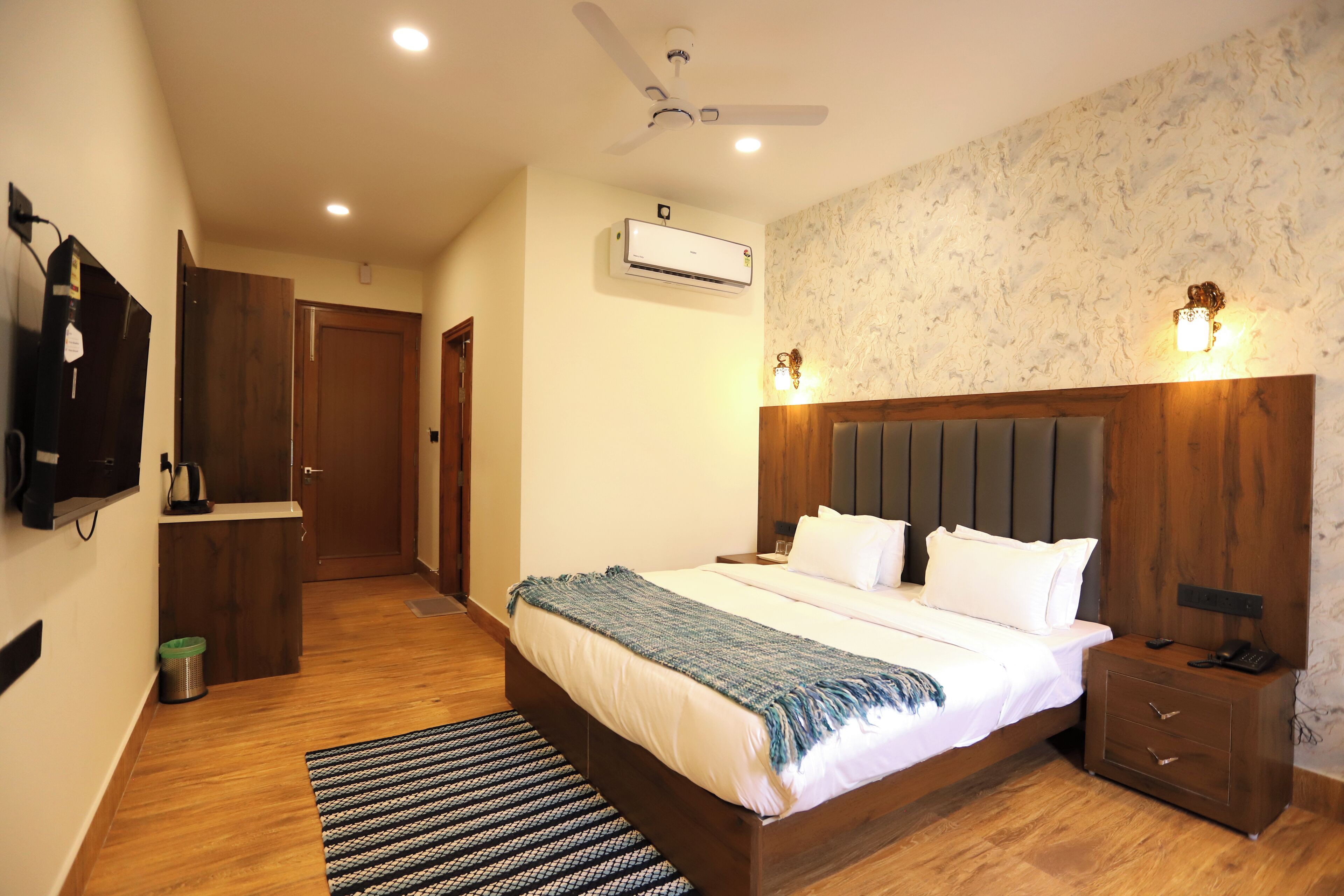 A bedroom with a wooden bed, a ceiling fan, an air conditioning unit, a bedside table with a lamp, and a small trash bin.