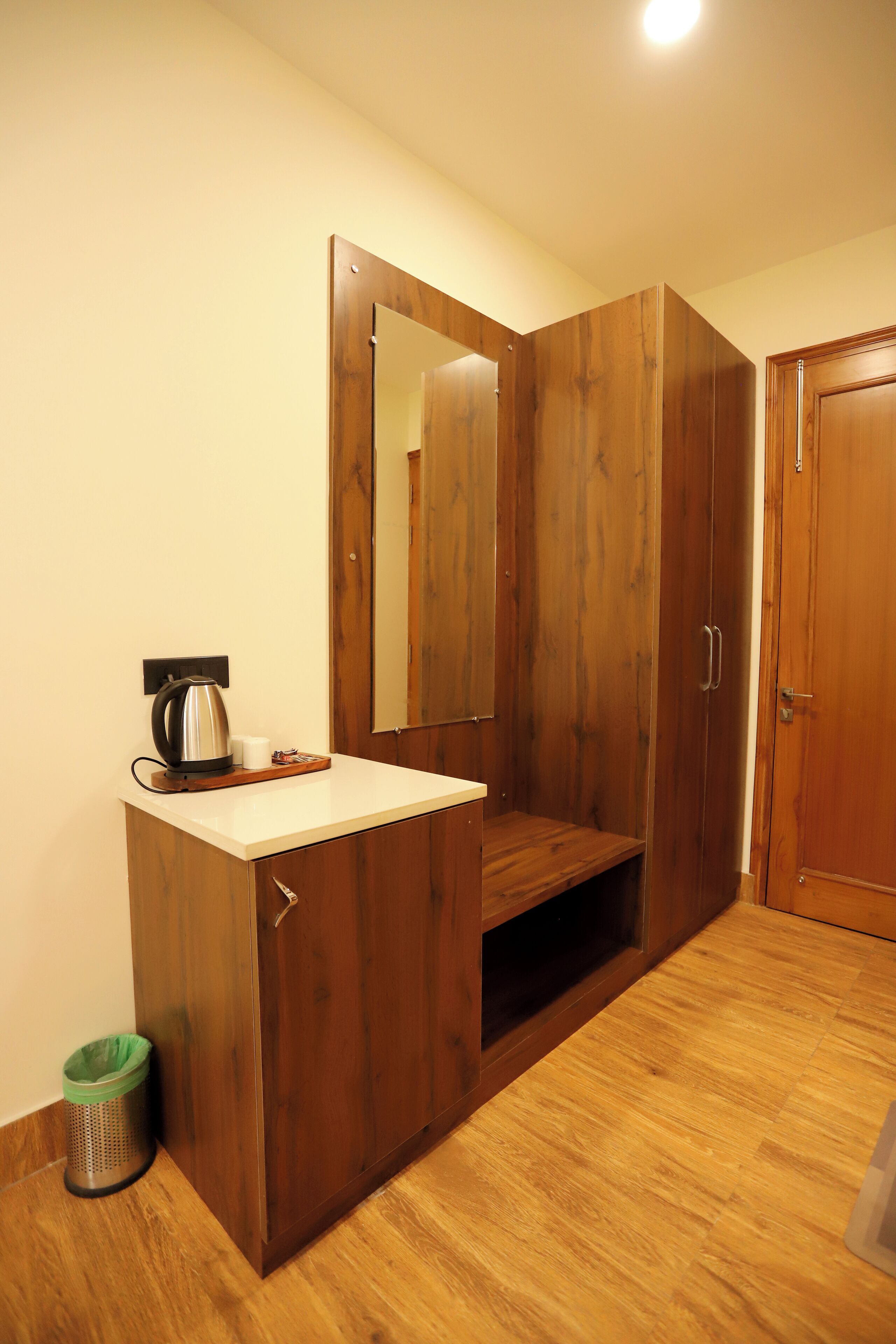 A wooden wardrobe with a mirror, a small table with a kettle and cup, and a trash bin.