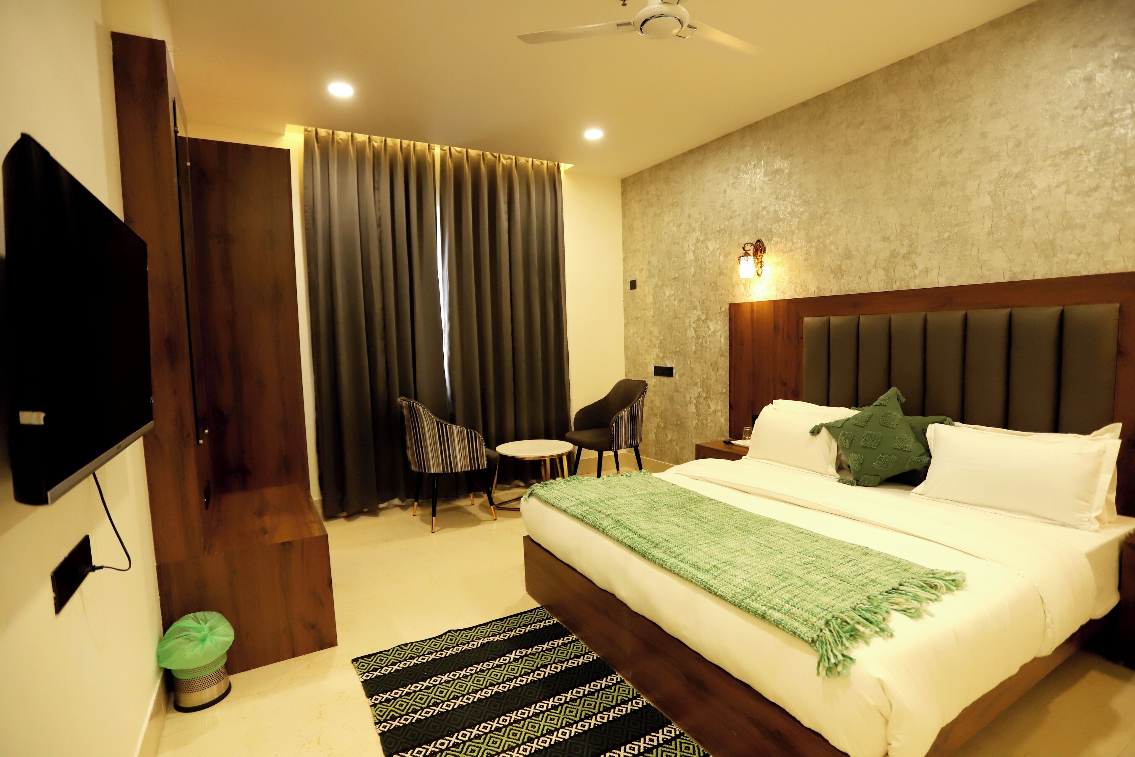 A hotel room with a large bed, a flat-screen TV, a small table, two chairs, a ceiling fan, and a window with curtains.