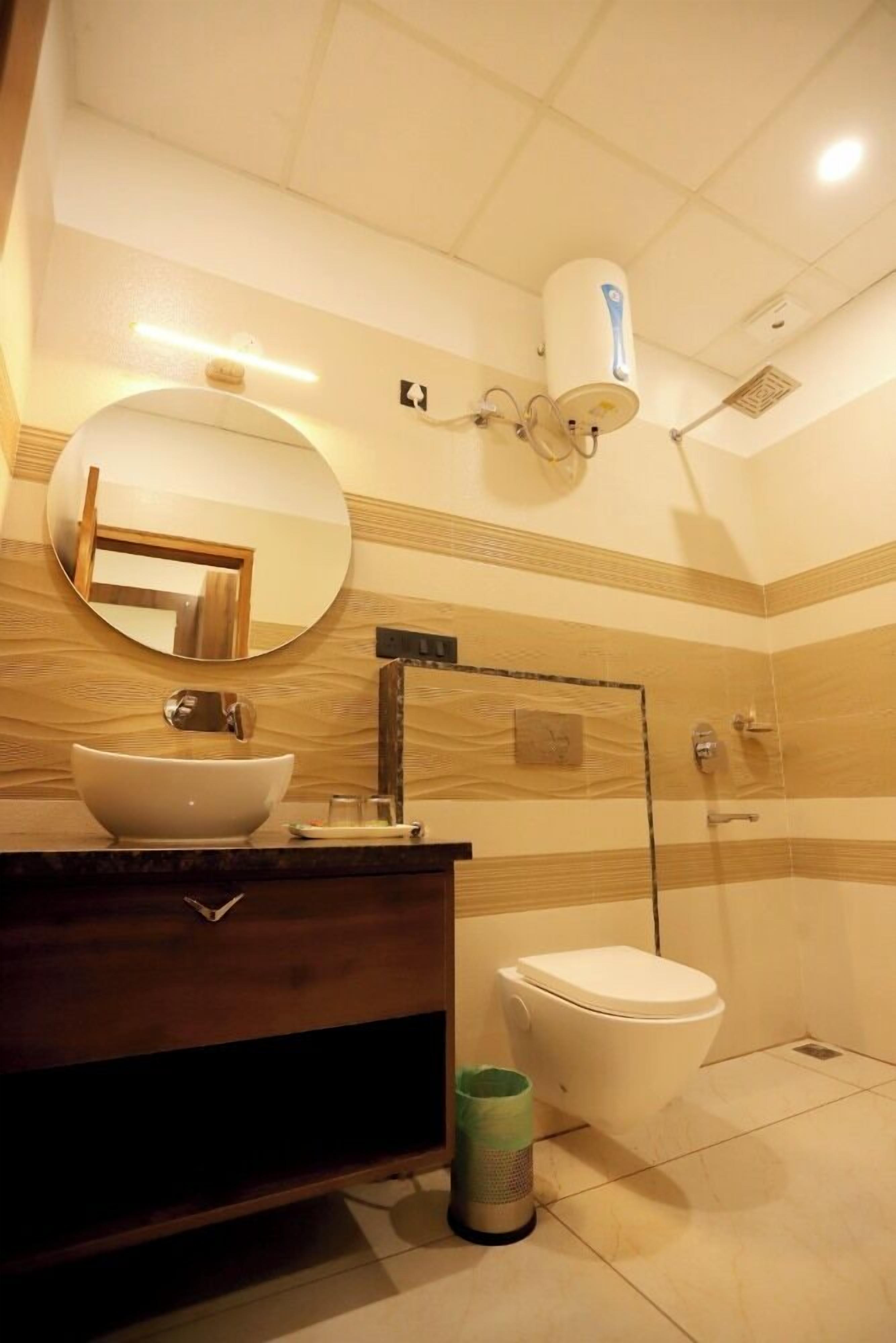 A bathroom with a wooden vanity, a round mirror, a white sink, a toilet, and a shower area.