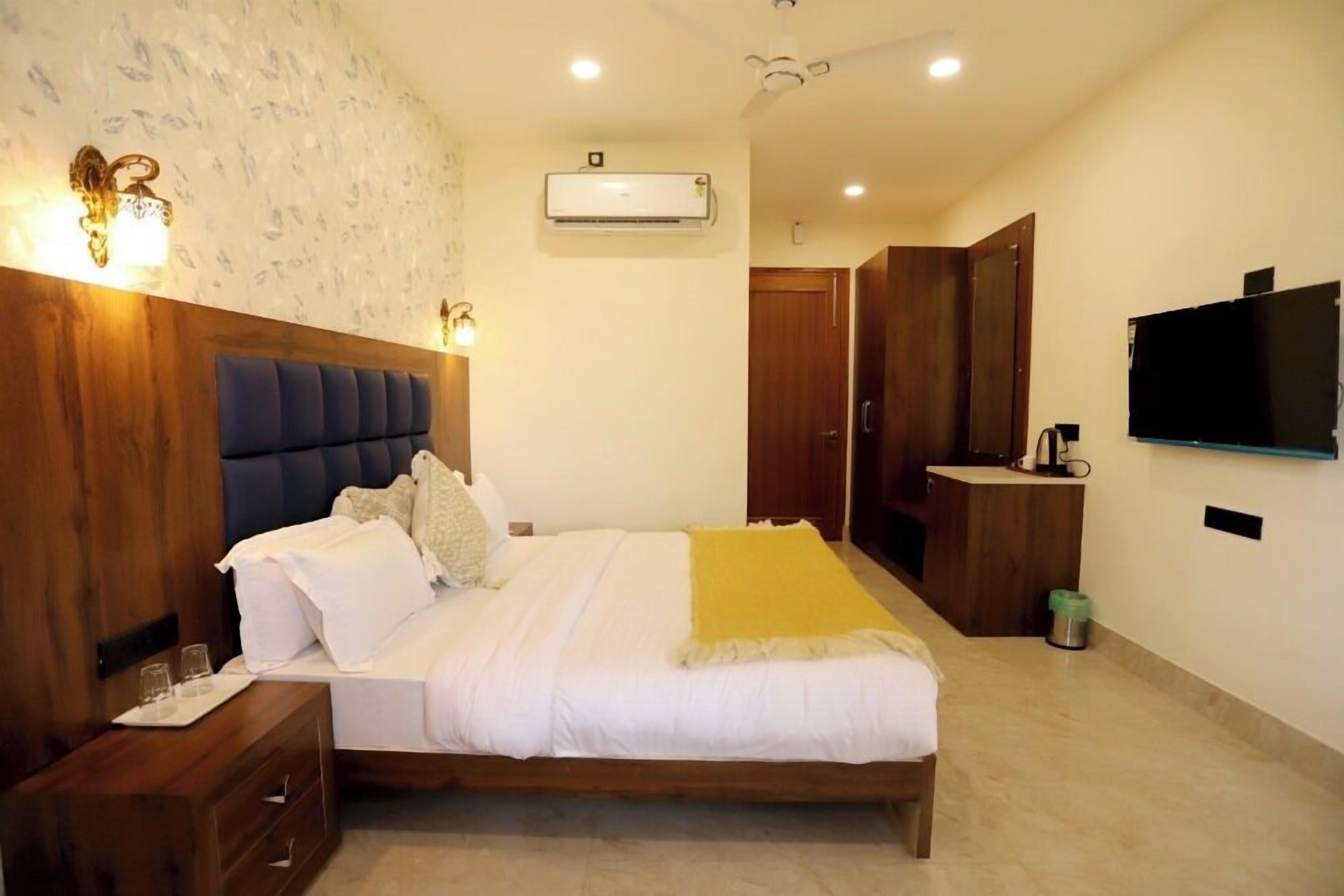 A bedroom with a bed, bedside tables, an air conditioning unit, a television, and a small kitchenette area.