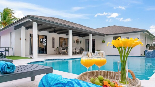 A poolside area with lounge chairs, a wicker basket with fruits and drinks, and a view of a modern house with a covered patio. - Grand Lux Villa /Heated Pool/Spa/Game fun/Nr Beach (Lantana)
