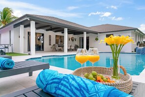 Zona de piscina amb gandules, una cistella de vimet plena de fruita i begudes, i vista cap a una casa moderna amb un porxo cobert.