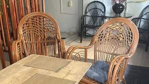 Een eetgedeelte met rieten stoelen en een geweven tafel, omringd door een grijs vloerkleed en een houten scheiding.