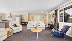 A spacious living area with a sofa, armchair, and coffee table, a kitchen in the background, and a large window with curtains. - Perfect Cromwell Family Getaway! (Cromwell)