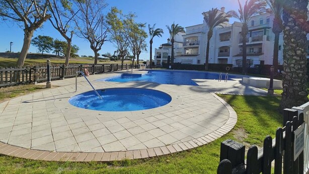 A swimming pool area with a circular design, surrounded by a paved walkway, palm trees, and a building in the background. - Comfortable 2 bed apartment - La Torre Golf AA1911 (Murcia)