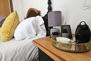 Una cama bien hecha con un almohadón blanco y un cojín decorativo amarillo, una mesita de noche con una lámpara, un altavoz negro y una cesta decorativa.