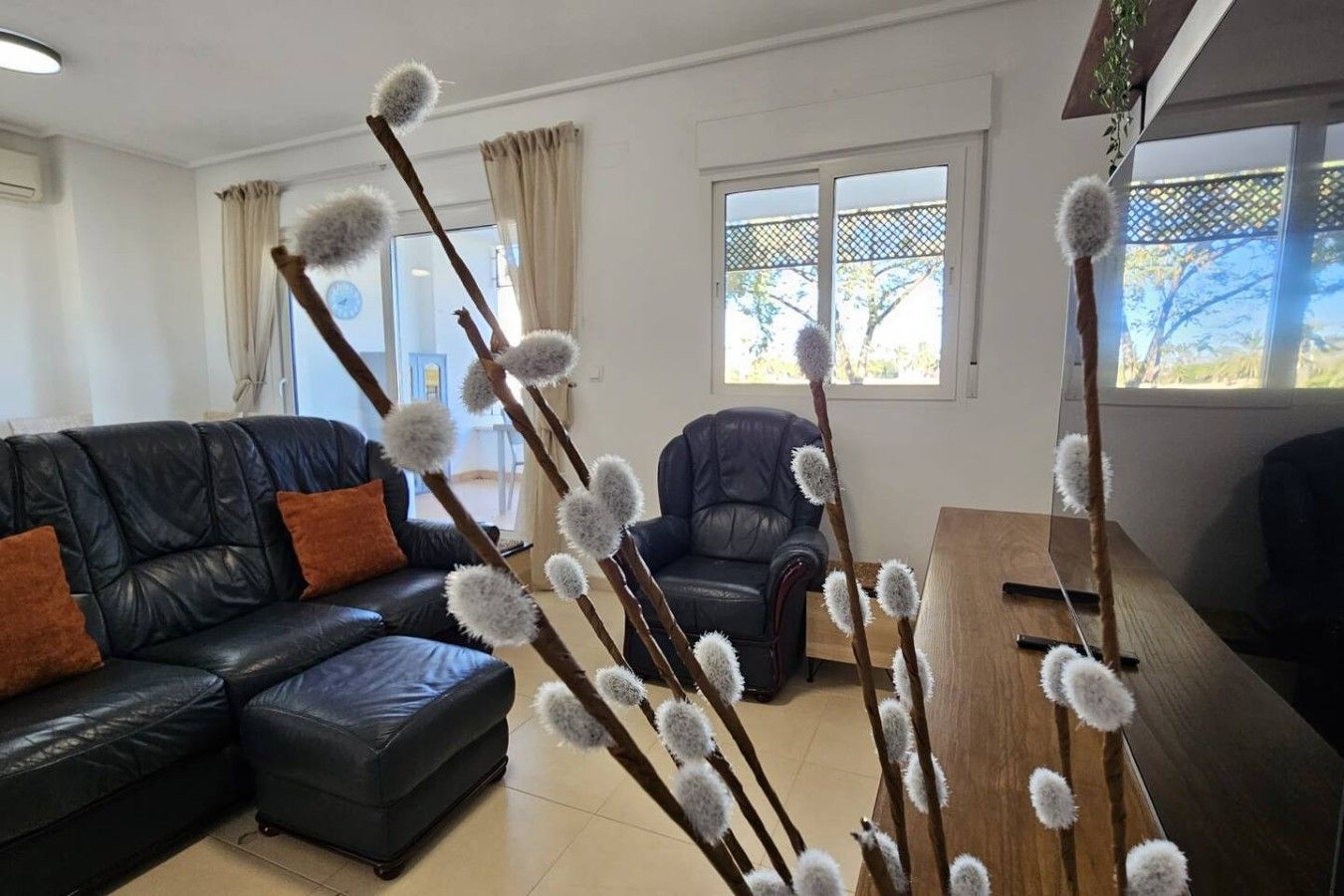 Een woonkamer met een zwarte leren bank, een houten salontafel en een fauteuil. Er staan decoratieve takken met witte, pluizige bolletjes.