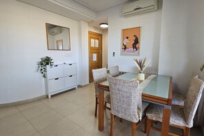Un área de comedor con una mesa de vidrio, sillas tejidas, un mueble blanco con cajones, una puerta de madera y un cuadro en la pared.
