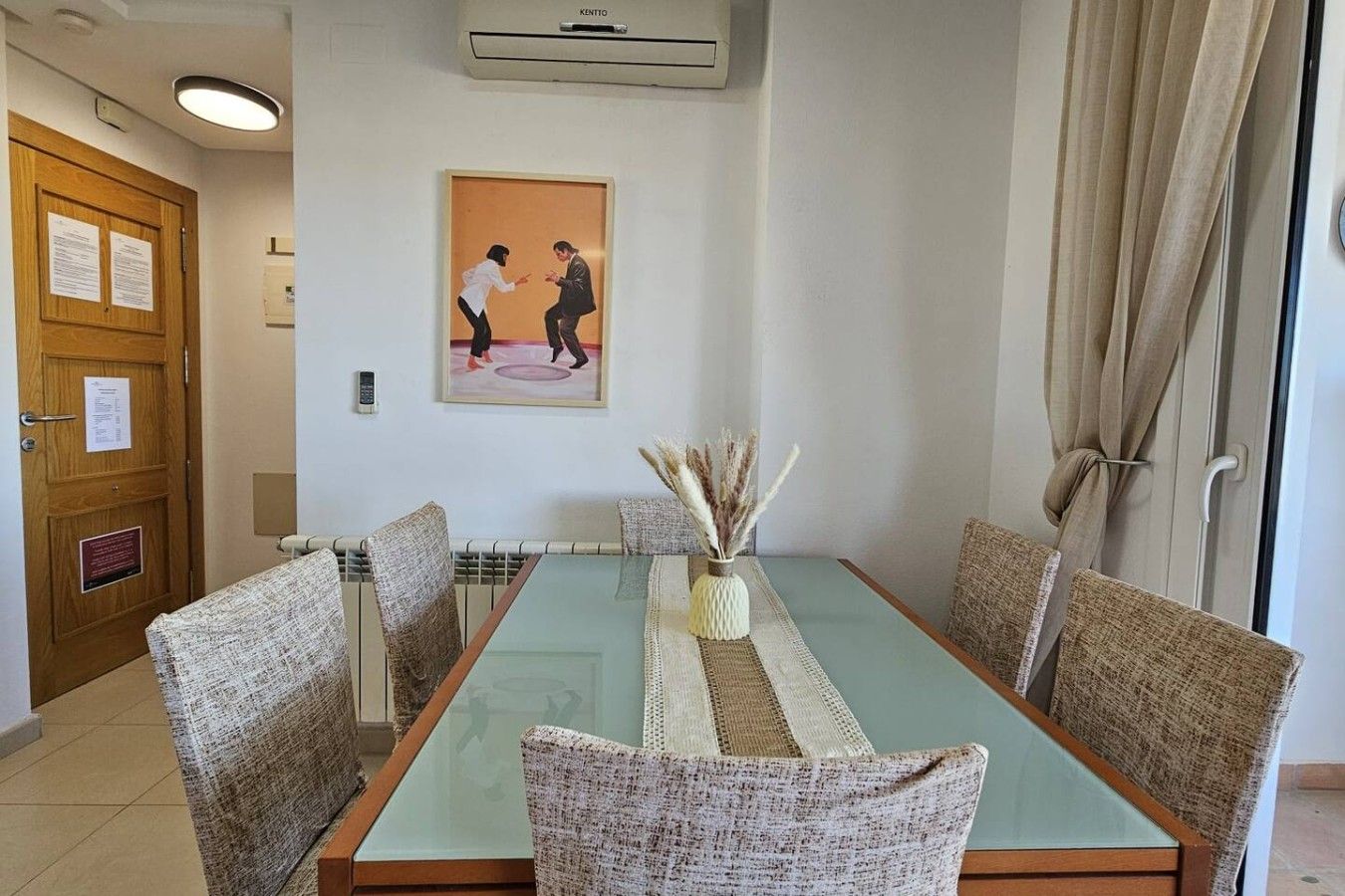 Un coin repas avec une table à plateau en verre, des chaises en osier, un vase contenant des plantes séchées et un tableau accroché au mur.