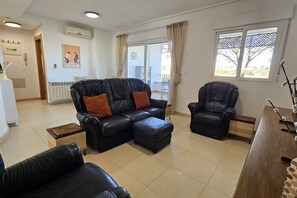 Ein Wohnzimmer mit einer Sofagarnitur, einem Sessel, einem Couchtisch und Blick ins Freie durch ein Fenster.