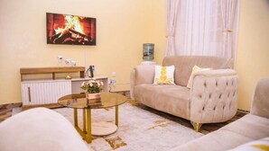 A living room with a beige sofa, a round glass coffee table, a white cabinet, and a flat-screen TV mounted on the wall. - Havana Cottages-Telaviv (Nakuru)