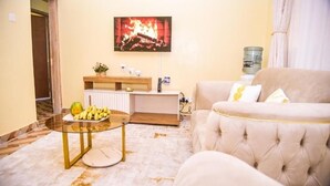 A living room with a beige sofa, a glass coffee table with fruit, a TV mounted above a fireplace, and a water dispenser on the wall. - Havana Cottages-Telaviv (Nakuru)