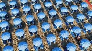 Une vue aérienne d’une plage où l’on aperçoit des rangées de parasols bleus et de chaises longues disposées selon un motif en quadrillage.