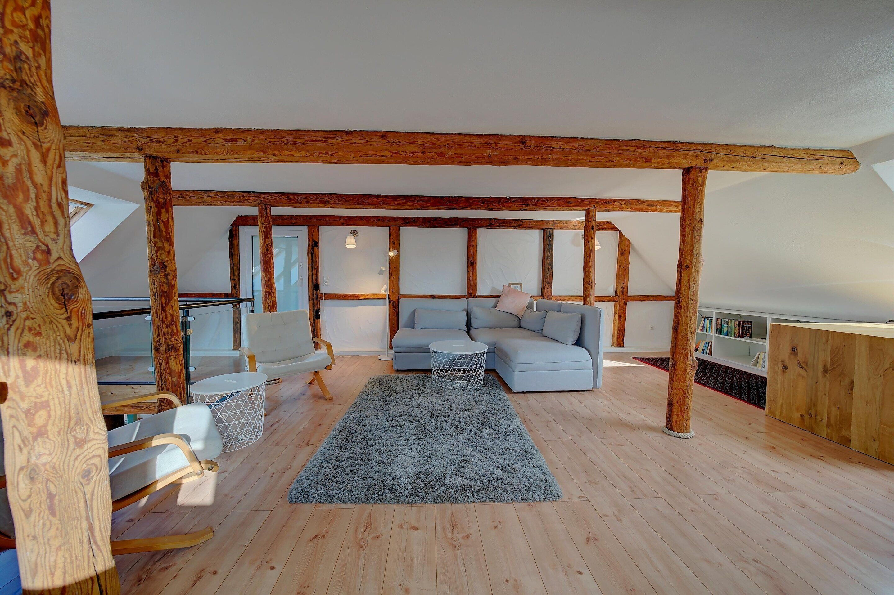 A spacious loft with exposed wooden beams, a gray sectional sofa, a white round coffee table, and a gray shag rug.