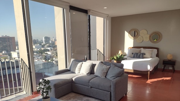 A modern living room with a grey sectional sofa, a white bed, and a large window offering a city view. - Luxury Urban Apartment in Chapultepec (Guadalajara)