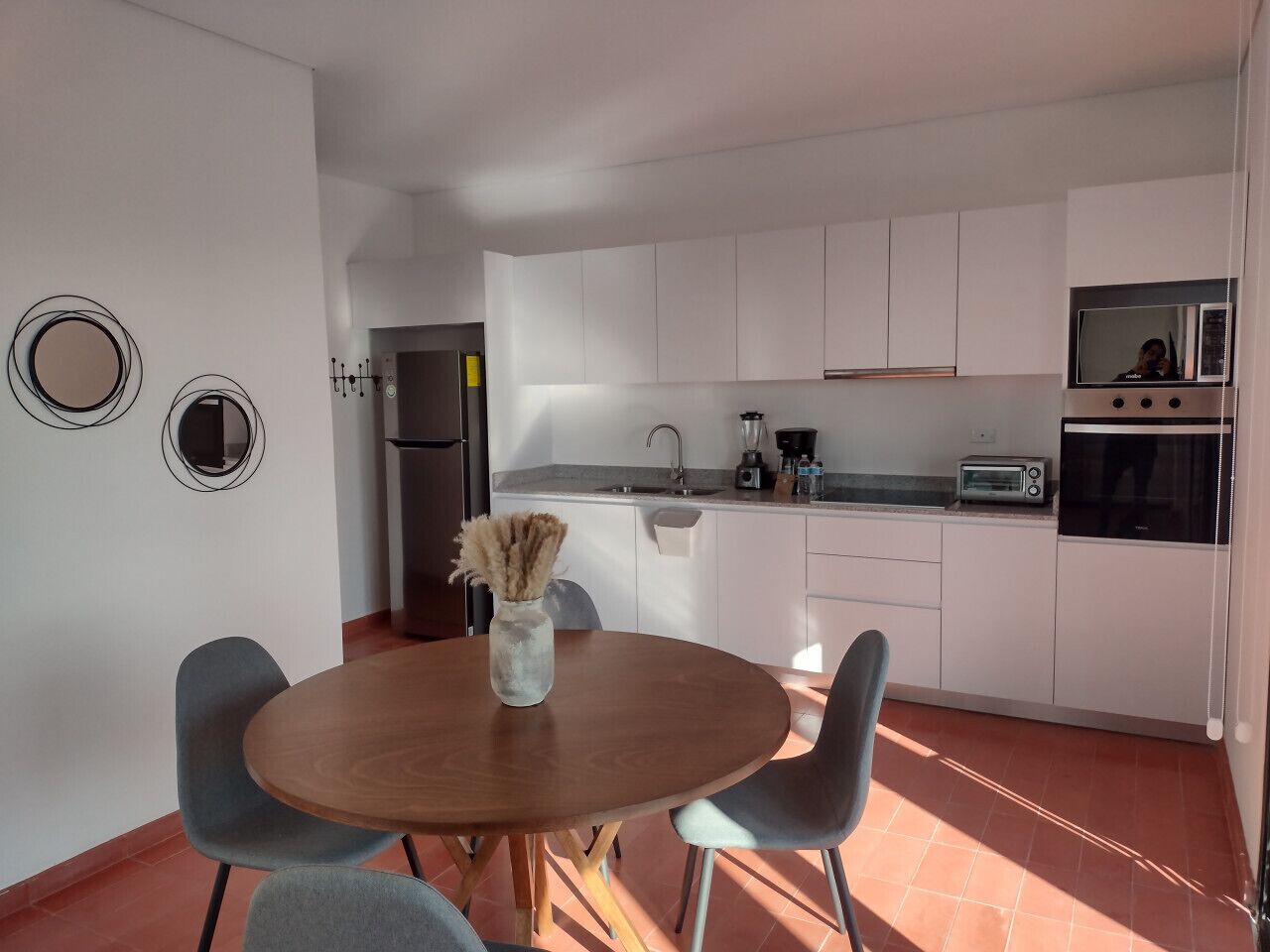 Une cuisine moderne avec des meubles blancs, un four encastré et un four à micro-ondes. On y trouve une table à manger avec deux chaises et un vase contenant des fleurs séchées.